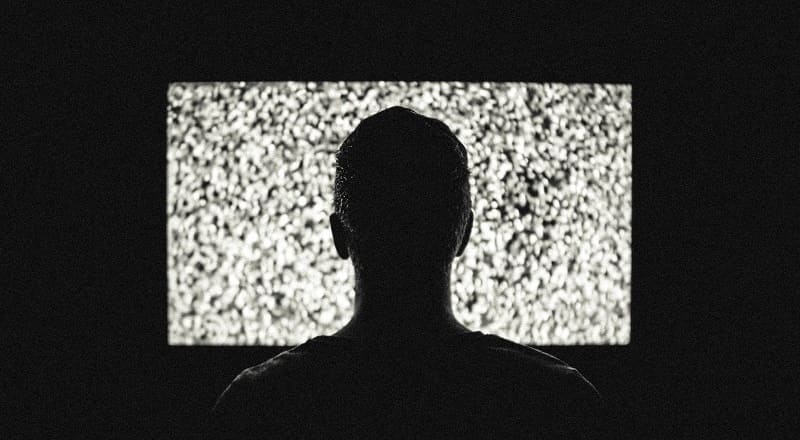 A man sitting in the dark in front of a TV screen full of static.